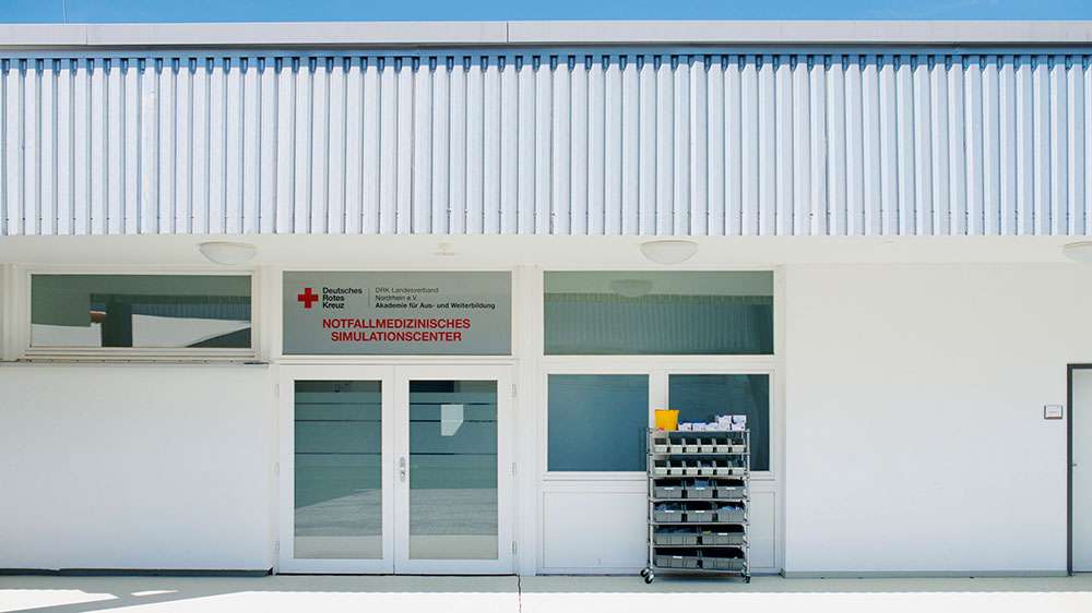 Eingang des Notfallmedizinischen Simulationscenters der DRK Akademie Nordrhein in Hennef mit Glasfront und rotem DRK-Kreuz über der Tür.